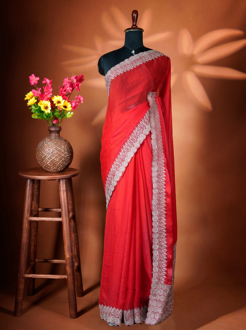 A red saree with a wide silver embroidered border is draped on a black mannequin torso. The mannequin is set against a warm brown background with decorative leaf shadows, and a small wooden stool holding a vase of colorful flowers is on the left.