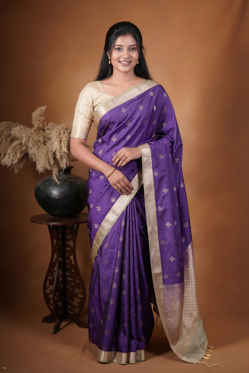 A woman modeling a purple silk saree with gold zari motifs and a beige blouse, standing against a warm brown studio backdrop next to a decorative vase.