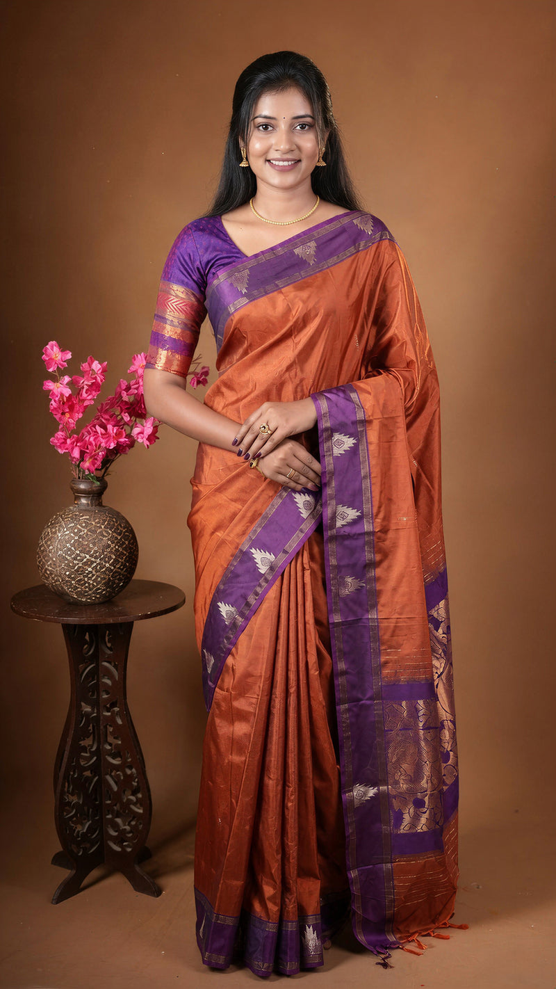 A woman modeling an orange and purple silk saree with gold zari work, standing next to a small table with pink flowers against a solid brown studio background.