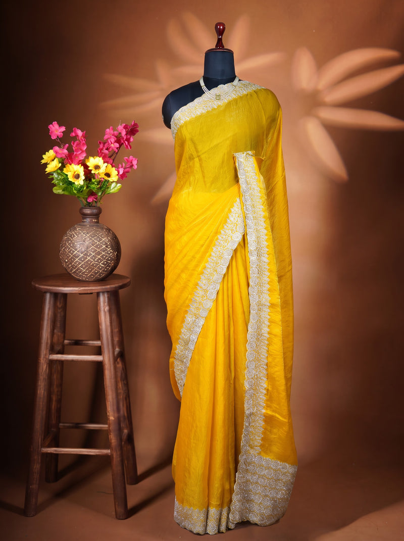 A bright yellow saree with a wide cream-colored embroidered border is draped on a black mannequin torso. The mannequin is set against a warm brown backdrop with decorative leaf shadows, and a small wooden stool holding a vase of colorful flowers is on the left.