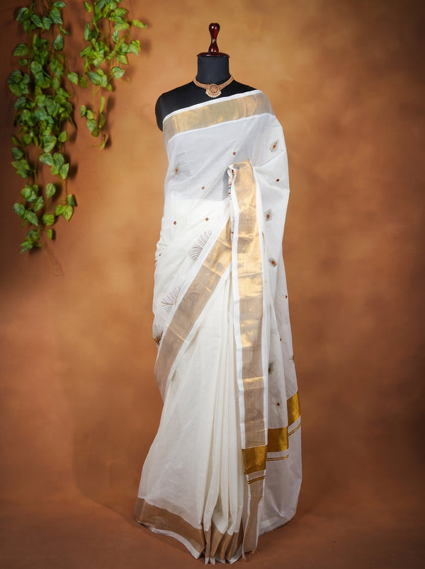 A white silk saree with a wide gold border and small gold motifs, displayed on a black mannequin torso against a warm brown backdrop with decorative green leaves on the left.