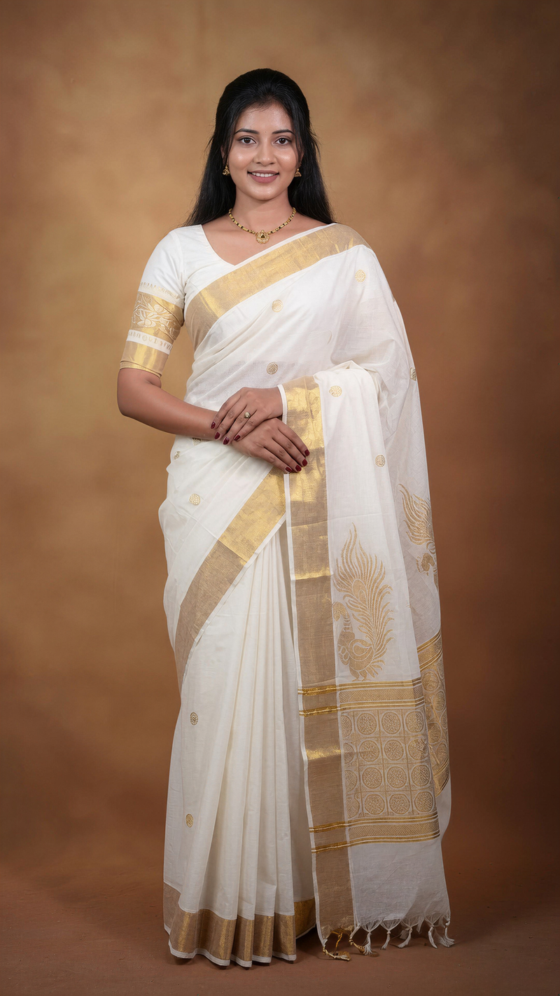 A woman modeling a white silk saree with a wide gold border and gold motifs, standing against a warm brown studio backdrop.