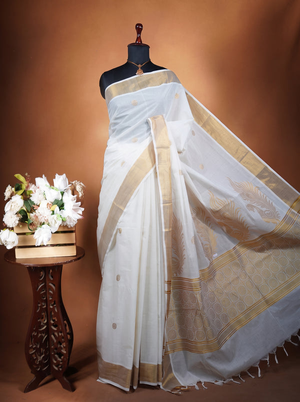 A white saree with gold borders and motifs displayed on a black mannequin torso, set against a warm brown studio backdrop. A small wooden table with white flowers is positioned to the left.