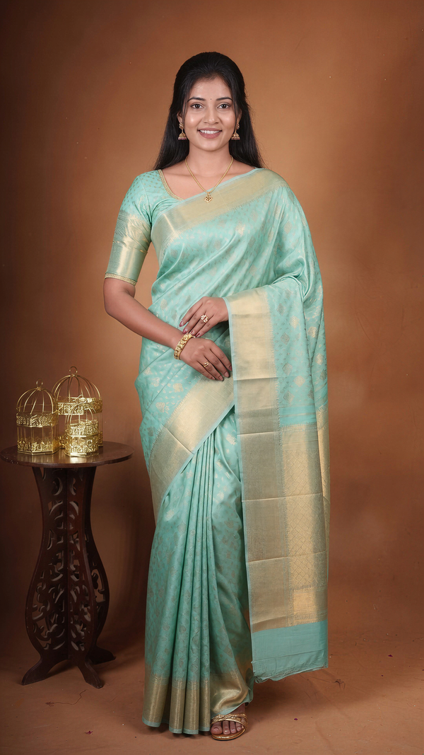 A woman modeling a light turquoise blue saree with a wide gold zari border and pallu, standing against a warm brown studio backdrop. She is posed next to a small dark wooden table holding two gold decorative lanterns.