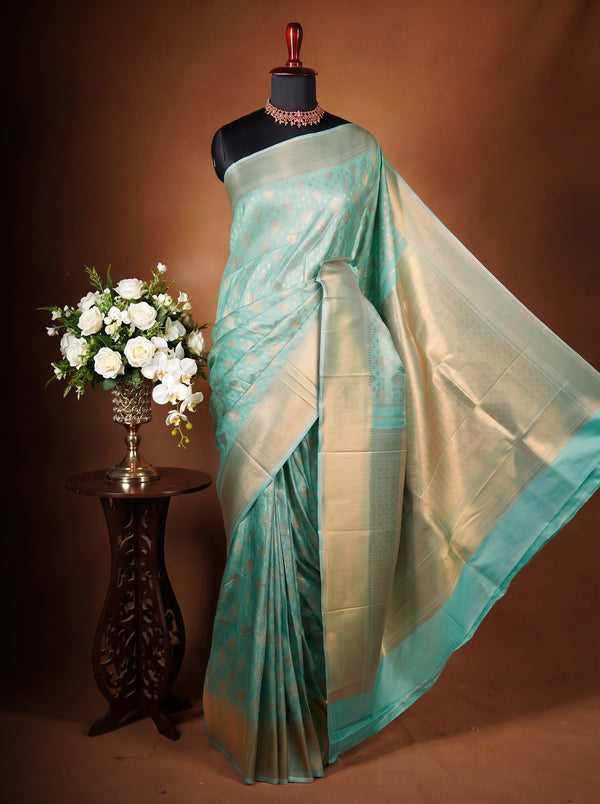 A light teal and gold zari silk saree draped on a black mannequin torso, styled with a decorative necklace, next to a small wooden table holding a white rose bouquet, against a warm brown studio backdrop.
