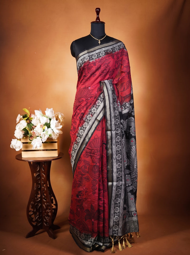 A red and black saree with intricate patterns and a silver border, displayed on a black mannequin torso. The mannequin is set against a warm brown studio backdrop, with a small wooden table holding white flowers on the left.