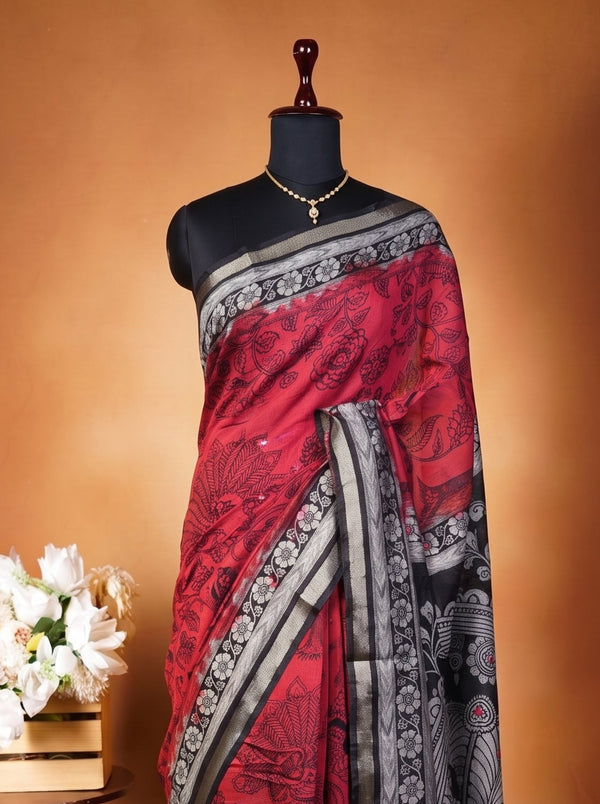 A red and black saree with intricate patterns and a silver border, displayed on a black mannequin torso. The mannequin is set against a warm brown studio backdrop, with a small wooden table holding white flowers on the left.