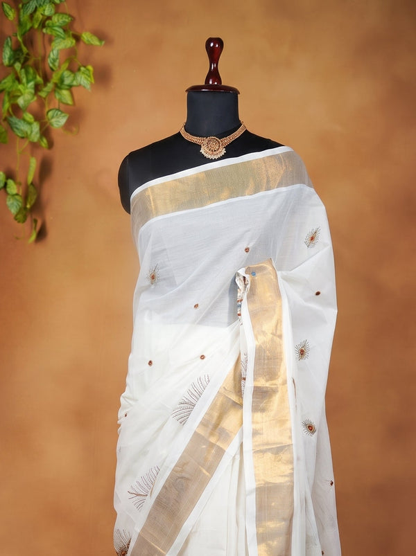 A white silk saree with a wide gold border and small gold motifs, displayed on a black mannequin torso against a warm brown backdrop with decorative green leaves on the left.