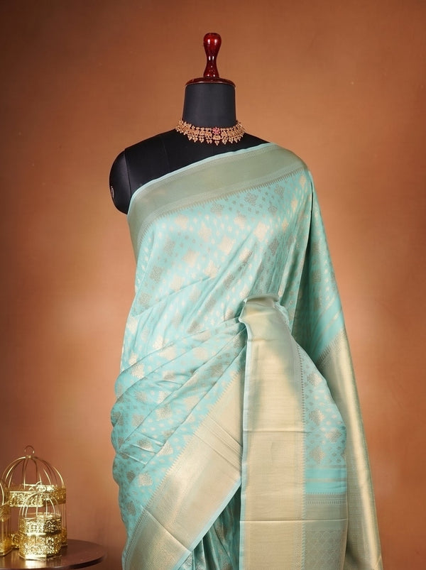 A light blue-green silk saree with gold zari motifs draped on a black mannequin torso, set against a warm brown studio backdrop with a small carved wooden side table holding two gold birdcages.