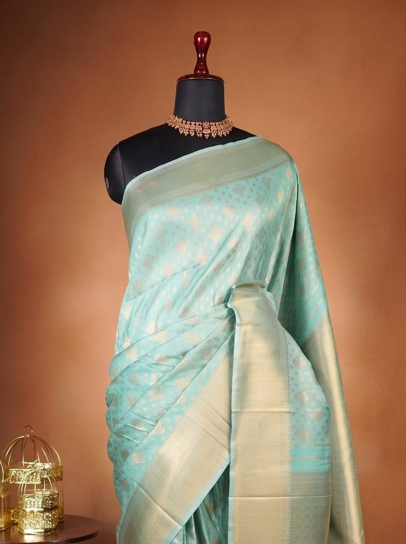 A light blue-green silk saree with gold zari motifs draped on a black mannequin torso, set against a warm brown studio backdrop with a small carved wooden side table holding two gold birdcages.