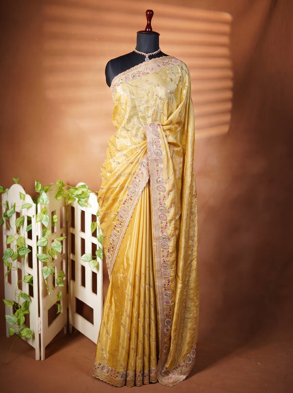 A mustard yellow silk saree with intricate silver and gold embroidery, draped on a black mannequin torso. The saree is styled with a matching embroidered border, set against a warm brown backdrop with a small white decorative screen on the left.