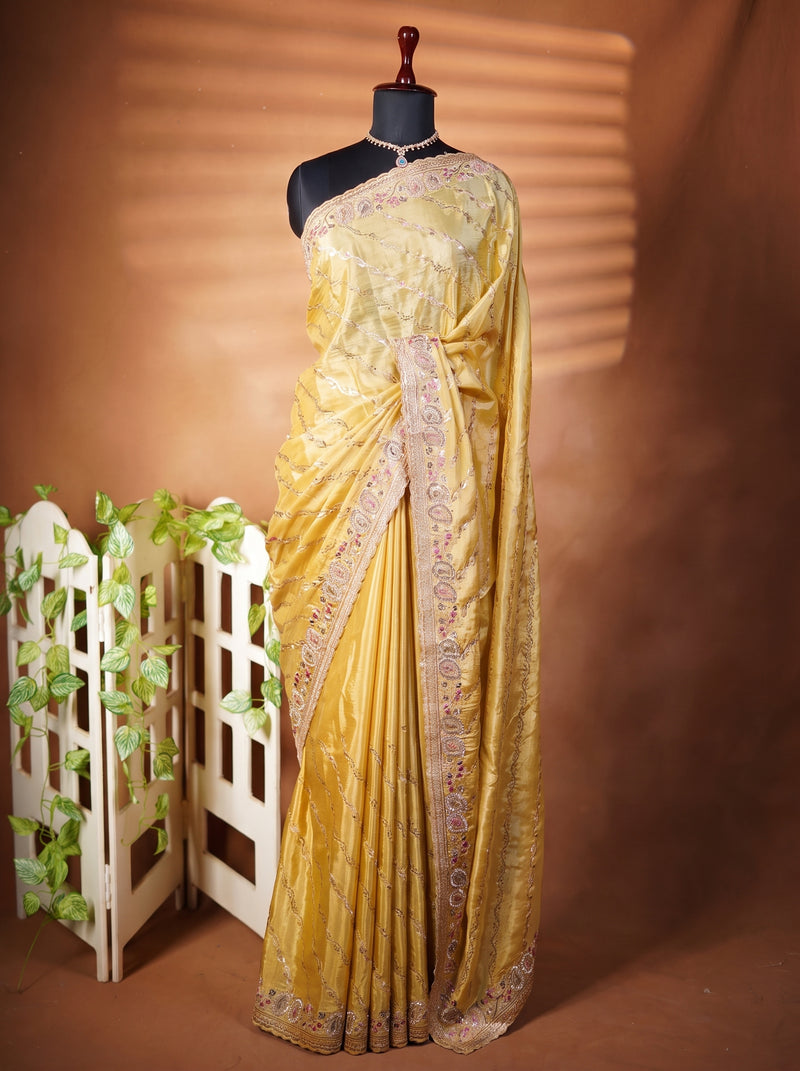 A mustard yellow silk saree with intricate silver and gold embroidery, draped on a black mannequin torso. The saree is styled with a matching embroidered border, set against a warm brown backdrop with a small white decorative screen on the left.
