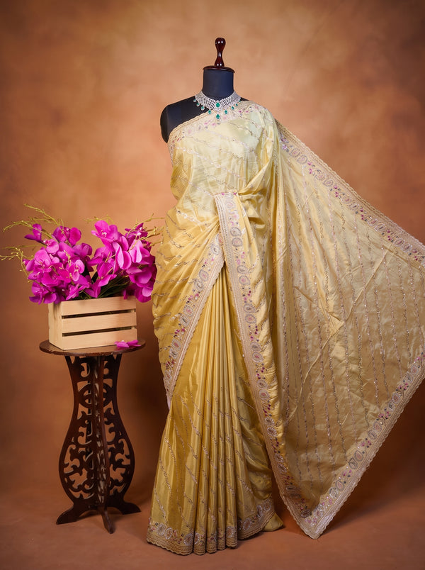 A light yellow silk saree with intricate floral embroidery on the border, displayed on a black mannequin. The saree is draped elegantly, paired with a matching embroidered blouse.
