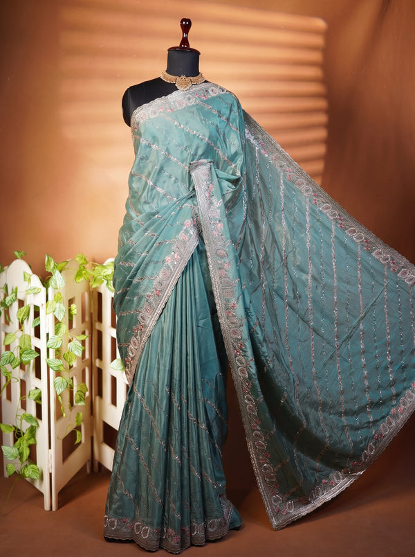 A teal green saree with silver floral embroidery and a matching border, displayed on a black mannequin against a warm brown backdrop with decorative plants.