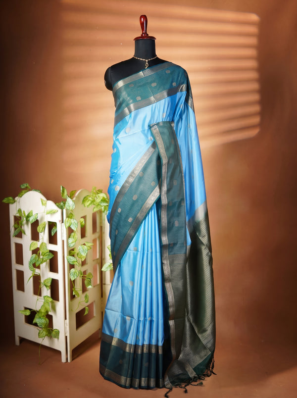 A blue and black saree with metallic gold borders displayed on a black mannequin torso, set against a warm brown background with decorative shadow bands and a small white plant stand.