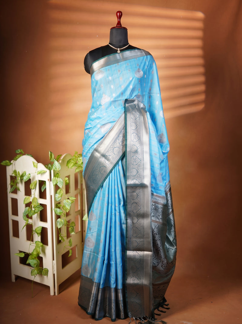 A light blue silk saree with a metallic silver border and pallu, draped on a black mannequin torso. The mannequin is set against a warm brown backdrop with a decorative white wooden screen and green vines on the left.