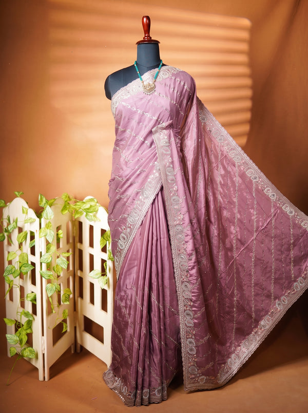 A mauve purple saree with silver zari embroidery draped on a black mannequin, styled with a turquoise necklace. The background is a warm brown studio backdrop with a white decorative screen and green leaves on the left.