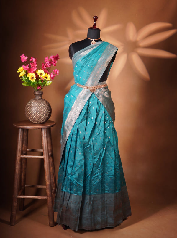 A teal blue and silver saree displayed on a black mannequin dress form, styled with a gold belt. The mannequin is set against a warm brown backdrop with decorative star-shaped shadows, and a rustic wooden stool holding a vase of colorful flowers is positioned to the left.