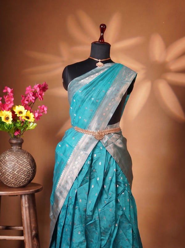 A teal blue and silver saree displayed on a black mannequin dress form, styled with a gold belt. The mannequin is set against a warm brown backdrop with decorative star-shaped shadows, and a rustic wooden stool holding a vase of colorful flowers is positioned to the left.