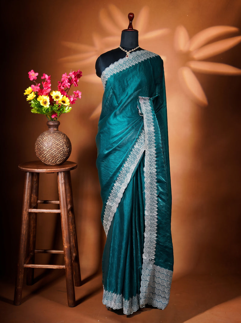 A teal green saree with a light gray and white floral lace border draped on a black mannequin torso. The mannequin is set against a warm brown background with leaf shadows, and a small wooden stool holding a vase of colorful flowers is on the left.