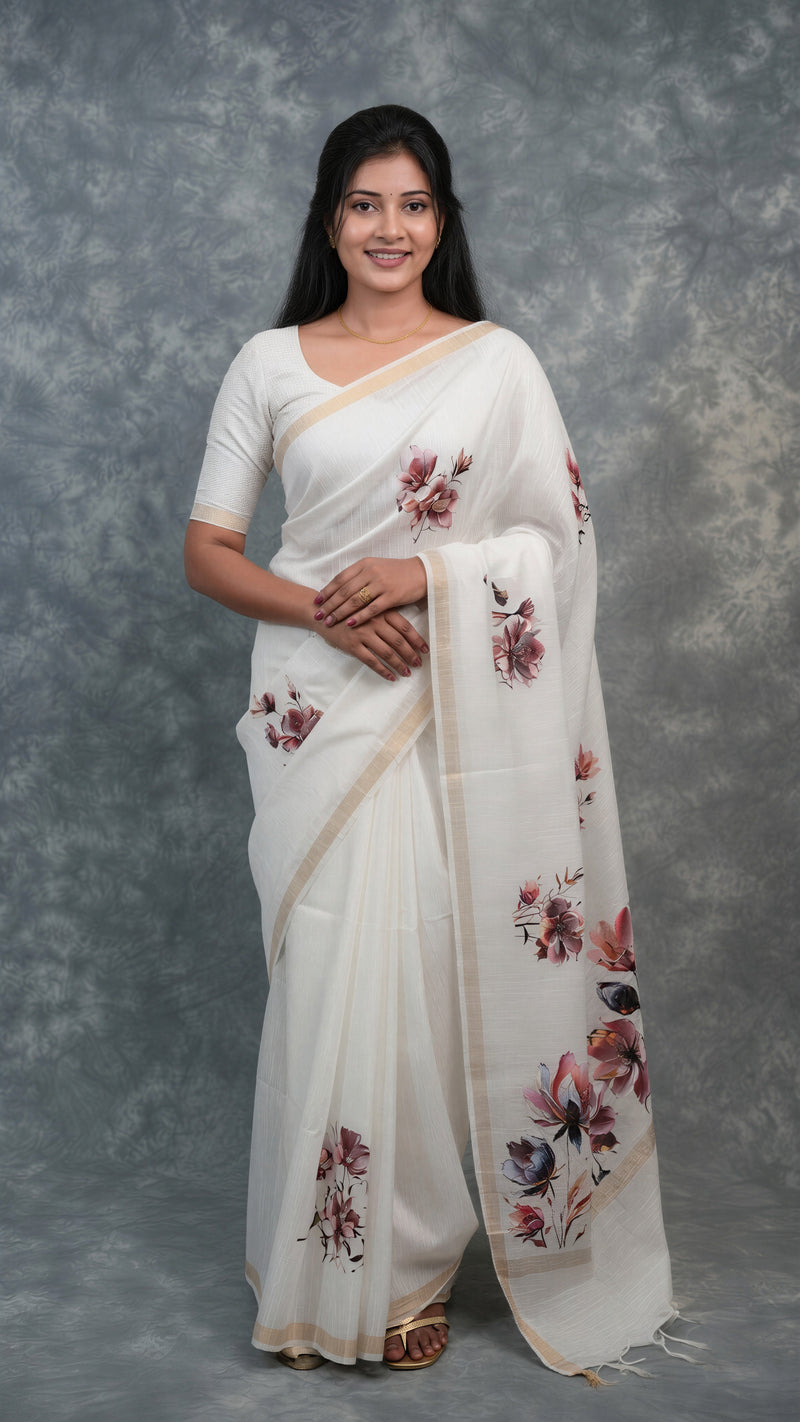 A woman modeling a white cotton saree with floral prints in pink, red, and green, paired with a matching white short-sleeve blouse, set against a textured gray studio backdrop.