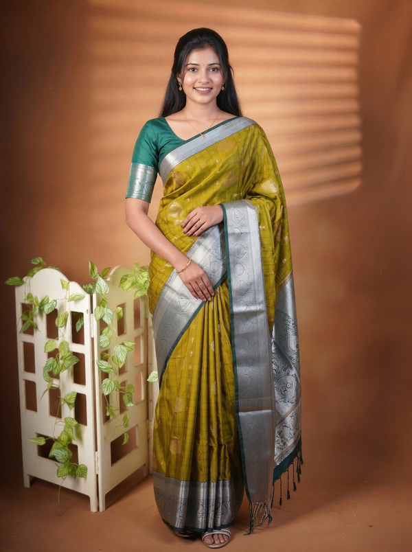 A woman modeling a traditional Indian saree with a mustard yellow body, a silver border, and a teal green blouse, standing against a warm brown studio backdrop with a decorative white screen and green vines.