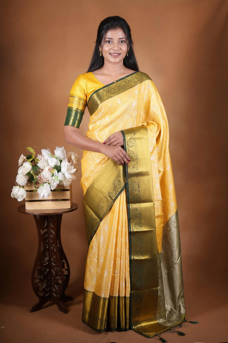 A woman modeling a yellow and gold zari woven saree with a green border, standing next to a small wooden table with a floral arrangement against a solid brown studio backdrop.