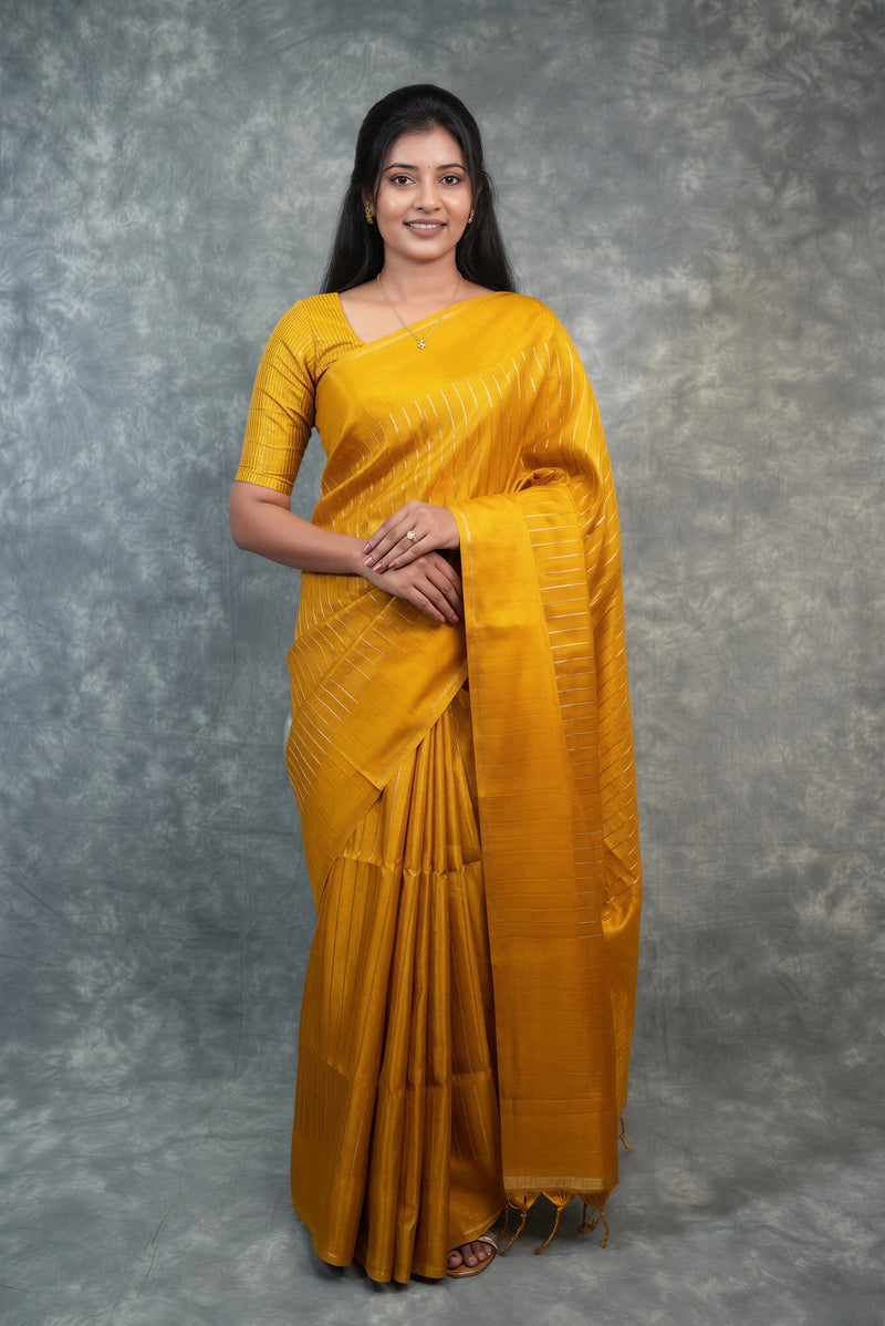 A woman modeling a mustard yellow silk saree with a matching short-sleeve blouse, standing against a textured gray studio backdrop.