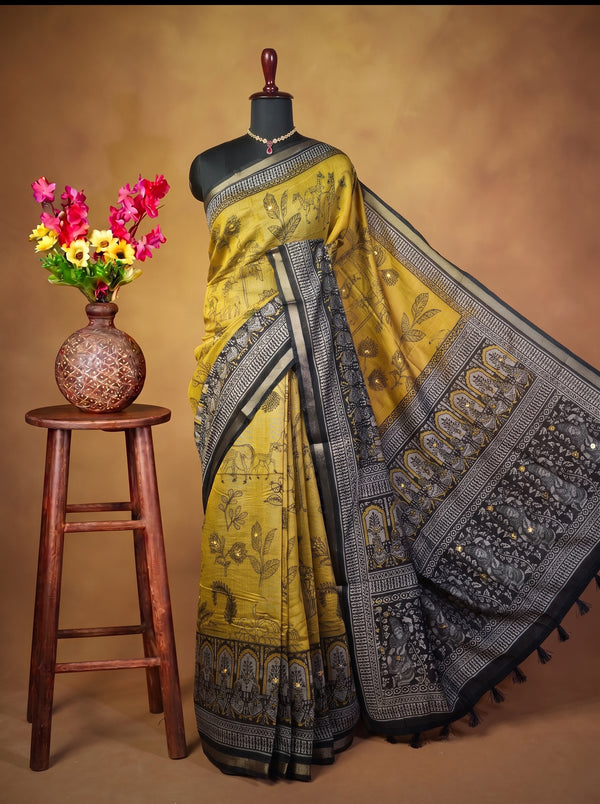 A mustard yellow and black saree with intricate floral and paisley motifs, displayed on a black mannequin. The saree is styled with a black border and is set against a warm brown studio backdrop, with a small wooden stool holding a vase of colorful flowers on the left.