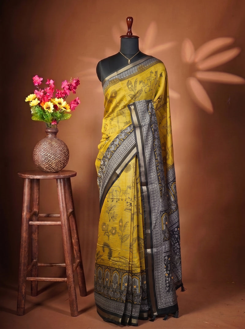 A yellow and black printed saree draped on a black mannequin torso, styled in a studio setting with a wooden stool holding a vase of pink and yellow flowers against a warm brown backdrop.