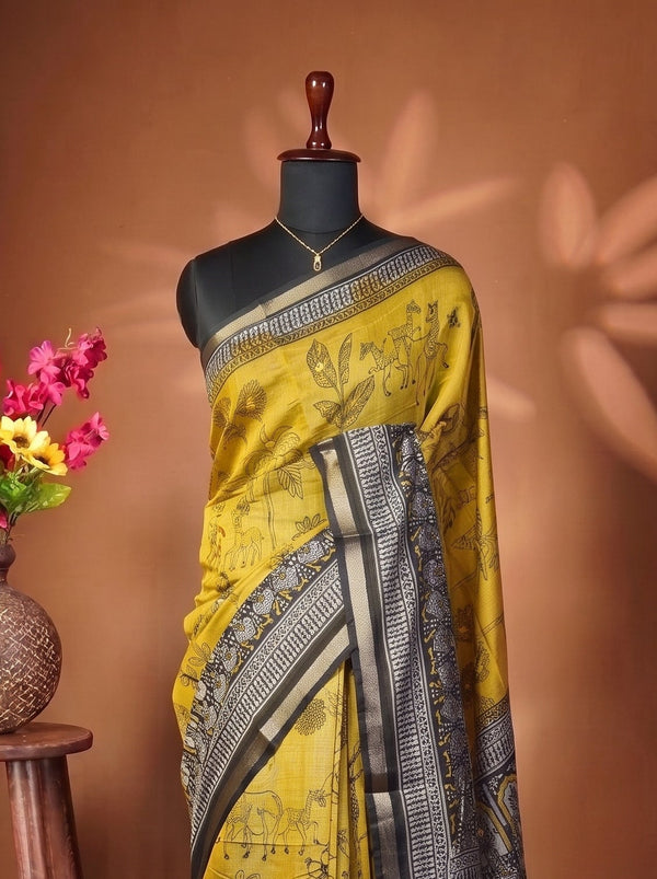 A yellow and black printed saree draped on a black mannequin torso, styled in a studio setting with a wooden stool holding a vase of pink and yellow flowers against a warm brown backdrop.