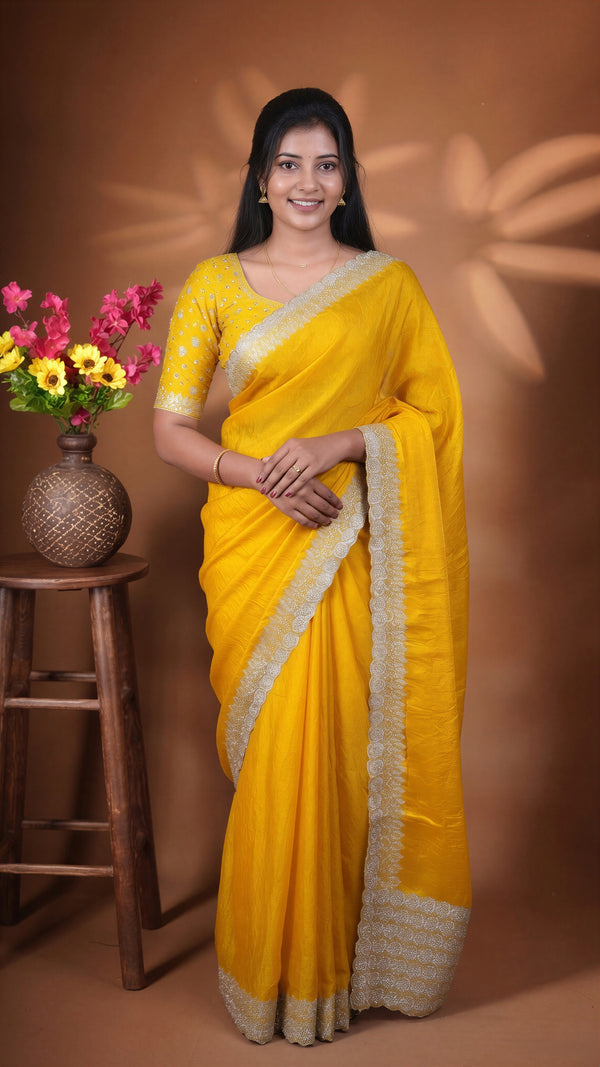A woman wearing a bright yellow sari with a beige border and matching blouse, standing in front of a brown studio backdrop with decorative shadows. A small wooden stool with a vase of flowers is visible on the left.