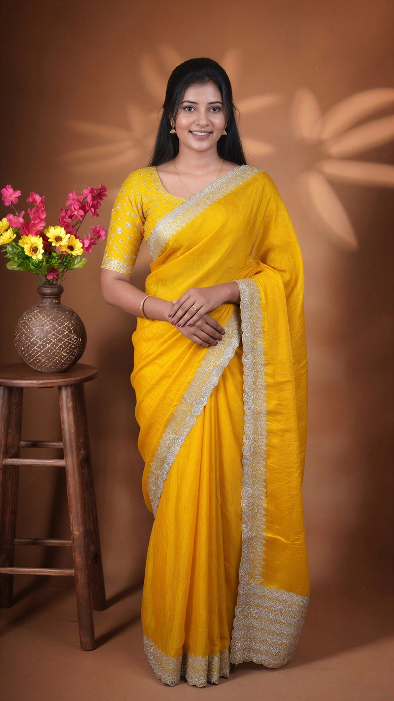 A woman wearing a bright yellow sari with a beige border and matching blouse, standing in front of a brown studio backdrop with decorative shadows. A small wooden stool with a vase of flowers is visible on the left.
