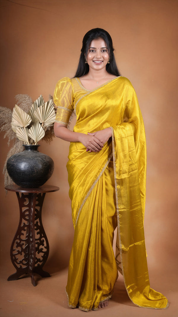 A woman wearing a bright yellow silk saree with a gold border and matching blouse, standing in front of a warm brown studio backdrop next to a decorative black vase on a carved wooden stand.